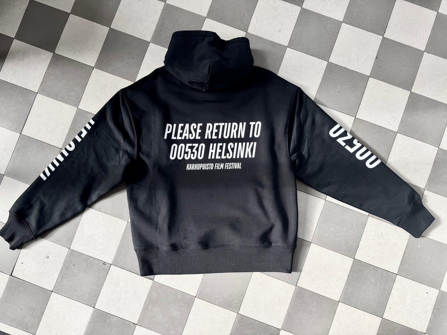 Karhupuisto Film Festival black unisex hoodie with white back print and sleeve details on a checkered floor