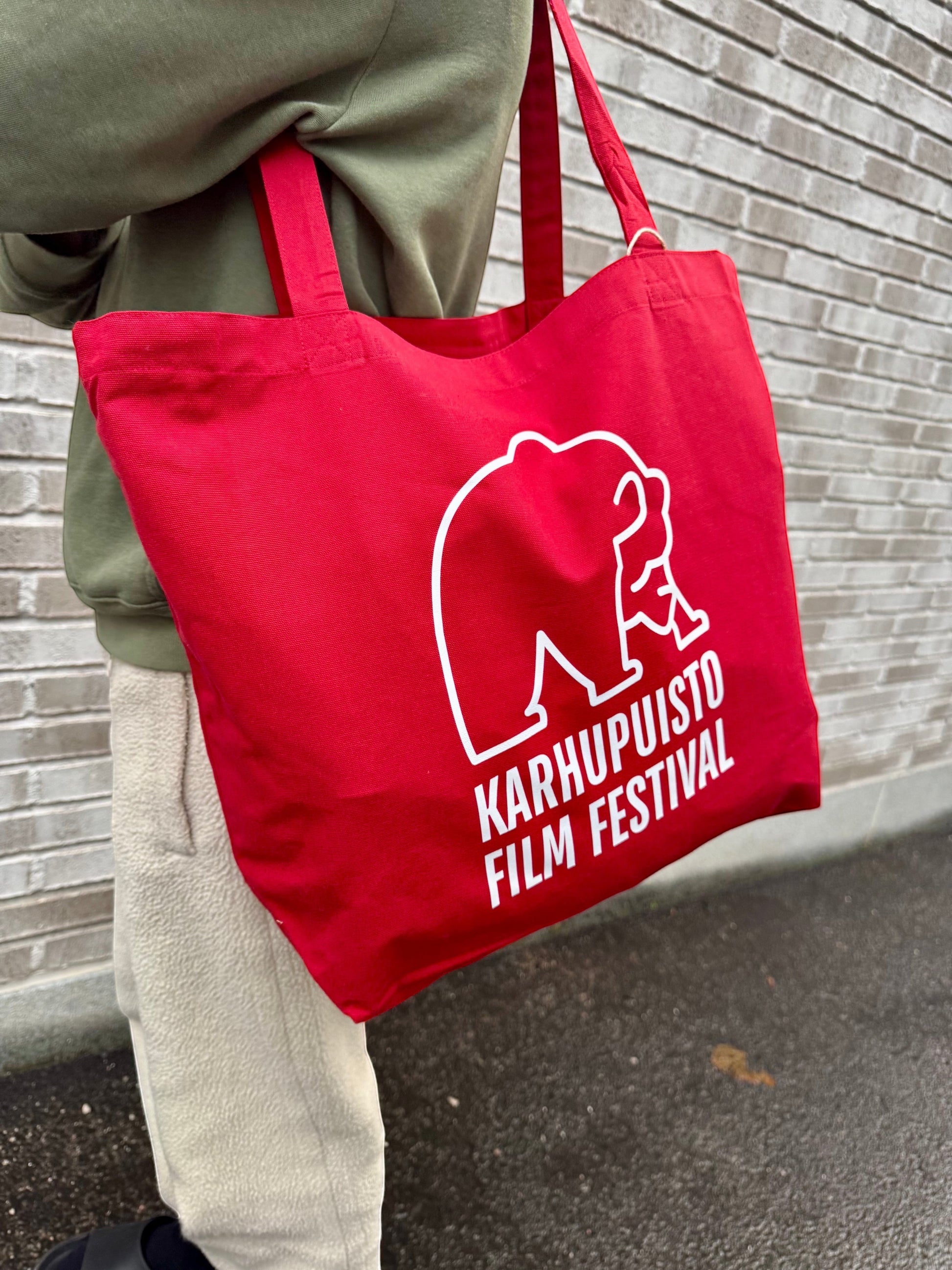 Karhupuisto Film Festival red cotton tote bag with white bear logo, worn over the shoulder against a brick wall.
