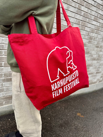 Karhupuisto Film Festival red cotton tote bag with white bear logo, worn over the shoulder against a brick wall.