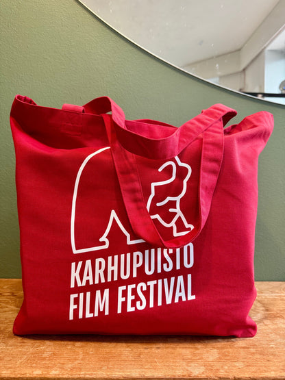 Karhupuisto Film Festival red cotton tote bag with white bear logo print on a wooden surface.