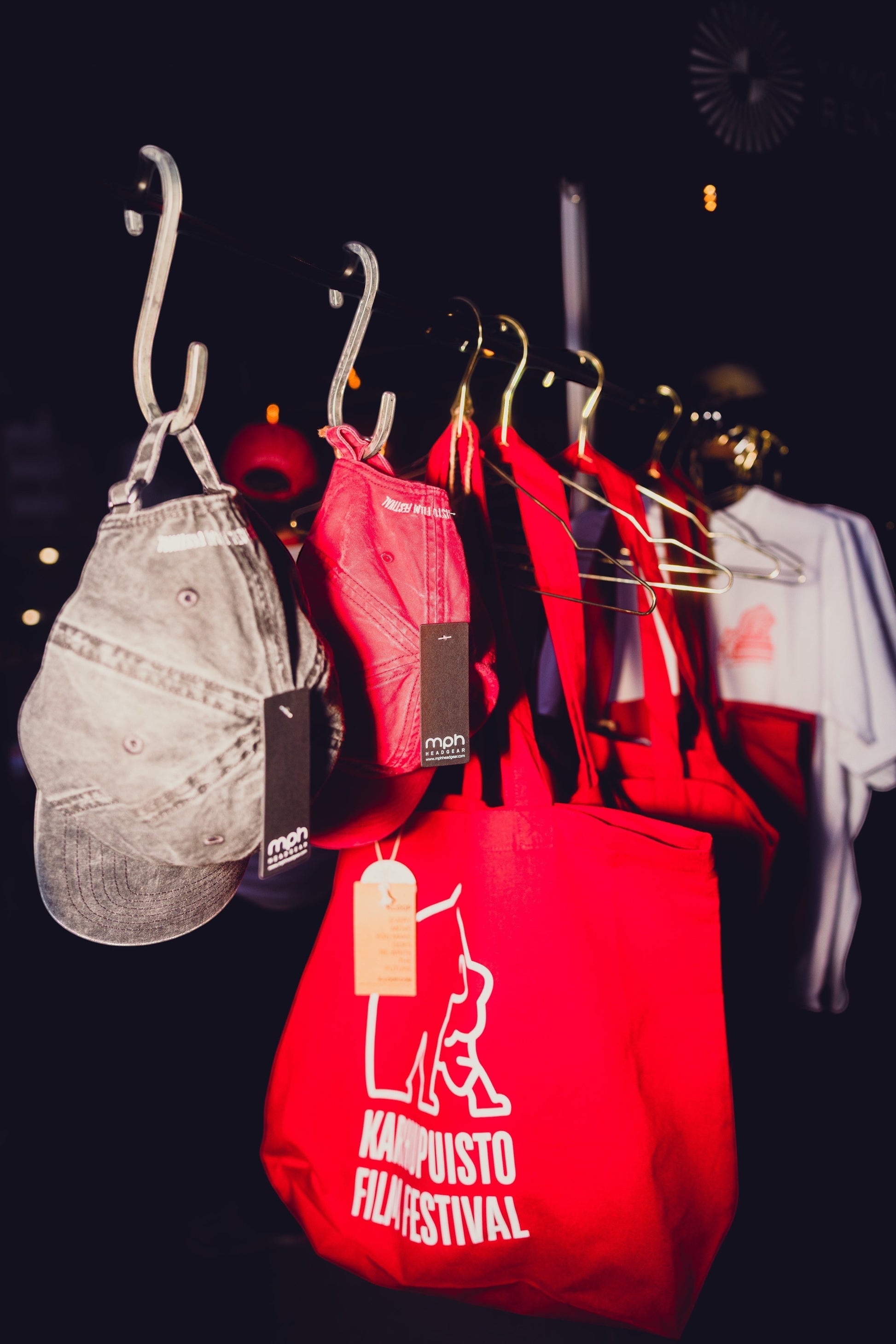 Karhupuisto Film Festival red cotton tote bag with white logo hanging on a rack with caps