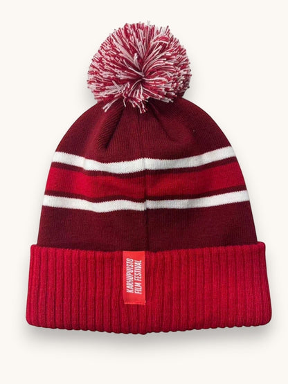 Karhupuisto Film Festival red and burgundy knit pom-pom beanie with white stripes and a ribbed cuff label.